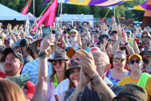Crowd at an outdoor festival with colorful banners overhead, people raising hands and cheering.