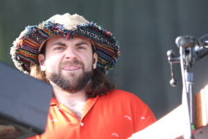 Man wearing a colorful woven hat and orange shirt singing into a microphone on stage