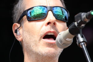 Close-up of a man singing into a microphone, wearing dark sunglasses that reflect a crowd and stage lights.