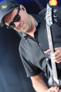 Guitarist wearing a dark cap and sunglasses, playing an electric guitar on stage with a strap over his shoulder.