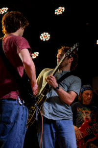 Three guitarists perform on stage under colorful overhead lights, captured from a close-up angle.