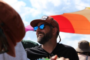 Man wearing a brown cap and reflective blue sunglasses smiles at an outdoor event, with colorful umbrellas in the bright sky behind him.