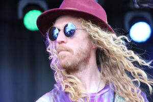 Close-up of a man with long blonde curly hair wearing a maroon hat and round mirrored sunglasses on stage.