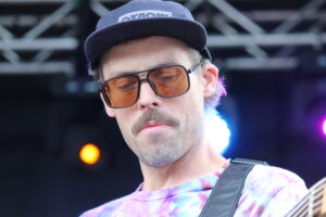 Male musician wearing a navy cap and orange-tinted sunglasses, playing a guitar on stage with colorful lights in the background.