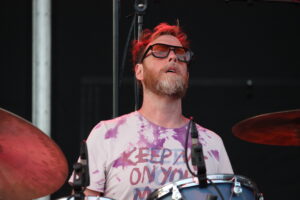 Drummer with red hair and a beard wearing orange-tinted glasses, playing a drum set on stage in a pink tie-dye shirt.