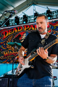 Male guitarist performing on stage with a sunburst electric guitar and colorful backdrop saying 'Deadpan' behind him, wearing a black shirt and jeans.