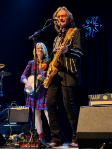 Man in a black suit plays a yellow guitar and sings into a mic on stage, with a woman playing banjo in a purple dress beside him.