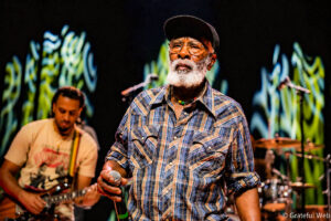 Older man with a white beard and glasses on a stage, wearing a plaid shirt and cap, holding a microphone while performing.