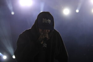 Hooded performer on stage, hat reads 'Dirty', holding a microphone under bright stage lights.