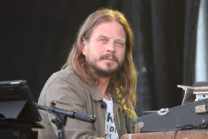 Bearded man with long hair sits at a keyboard on a dimly lit stage, wearing an olive jacket.