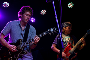 Two guitarists performing on a dark stage under colorful stage lights; left guitarist in a blue shirt plays a black electric guitar, right bassist with a bandana plays a red bass.