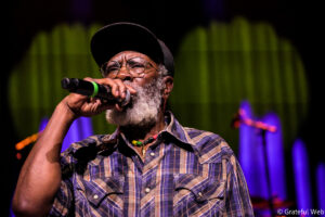 Male singer on stage wearing a plaid shirt and cap, holding a microphone