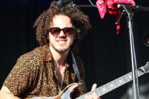 Musician with curly hair wearing sunglasses, playing an electric guitar on stage, patterned shirt visible.