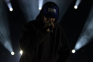 Performer wearing a dark hoodie and blue cap reading 'Dirty' singing into a microphone on a dimly lit stage with bright spotlights behind.