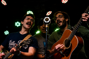 Three male musicians on a dim stage, two with acoustic guitars and one singing into a central mic under colored stage lights, guitar straps visible. (informative)