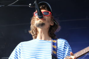 Musician wearing red sunglasses sings into a microphone on stage, blue-and-white striped shirt visible.