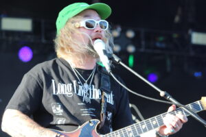 Male musician with long blond hair, green cap and white sunglasses singing into a microphone while playing an electric guitar on stage.