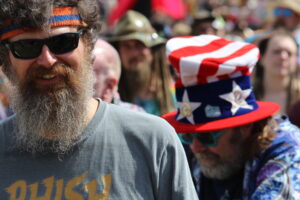 Bearded man wearing black sunglasses and an orange-blue headband at a crowded outdoor festival.