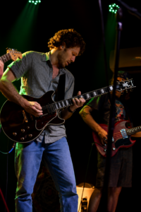 Guitarist in a striped shirt playing a dark electric guitar on a dimly lit stage with green spotlights.