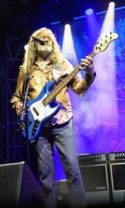 Male musician with long gray hair and beard playing a blue bass guitar on a concert stage with colorful stage lights behind him, singing into a microphone.