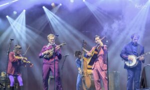 A group of musicians on stage wearing purple suits, playing string instruments under bright blue stage lights.