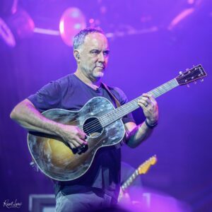 Male guitarist playing a sunburst acoustic guitar on stage under purple lights, wearing a dark shirt.