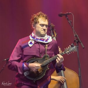 Male musician in a purple shirt and patterned scarf plays a mandolin on stage, with a microphone nearby.
