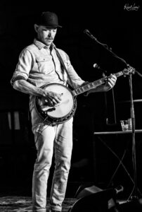 Male musician on a dark stage playing a banjo, wearing an NY cap and denim shirt, with a microphone nearby.