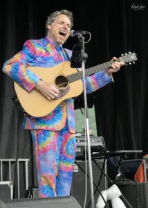 Man in a bright tie-dye suit passionately sings into a microphone while playing an acoustic guitar on stage.