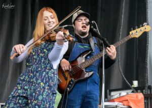 Two musicians perform on stage: a woman in a floral dress plays violin while a man in overalls and a cap sings into a microphone and plays bass guitar.