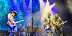 Three musicians perform on a illuminated stage: left guitarist in a wide-brimmed hat plays a blue electric guitar while two other guitarists sing and play, colorful backdrop behind them.
