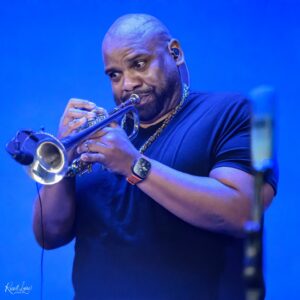 Close-up of a trumpet player in a black shirt, playing a silver trumpet under blue stage lights.