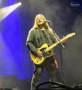 Male guitarist with long hair performing on stage, singing into a microphone under colorful stage lights.