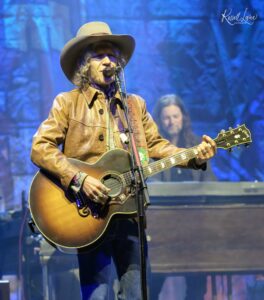 Guitarist wearing a tan cowboy hat and brown leather jacket performing on a blue-lit stage.