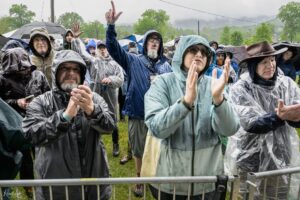 Group of people in rain ponchos and jackets gathered behind a metal barrier at an outdoor event in rainy weather, some holding cups or phones.