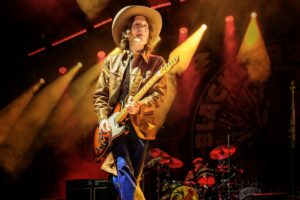 Guitarist in a cowboy hat and brown jacket playing an electric guitar on stage amid warm orange lights.