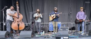 Four-member band performing on an outdoor stage: bassist left, two center guitarists/vocalists, and a right-side guitarist with a resonator guitar, with cables and gear on stage.