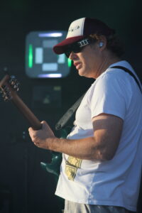 Male guitarist wearing a white T-shirt and red-trimmed cap, playing a green electric guitar on stage with in-ear monitors visible