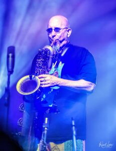 Male saxophonist performing on stage under blue lighting, wearing sunglasses and a black T-shirt.00The musician's horn reflected by vibrant stage lights.