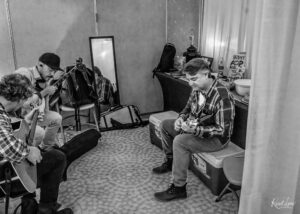 Two men seated in a backstage room, each playing an acoustic guitar among gear and bags, with a mirror propped against the wall behind them.