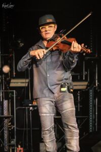 Violinist on stage wearing a cap that reads 'FIDDLE' and bright yellow glasses, playing a violin.