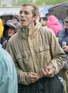 Young man in a tan rain jacket with damp hair, eyes closed, in a rainy outdoor crowd.