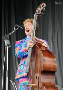 Singer in a bright tie-dye suit belts into a microphone while holding a large upright bass on stage against a black backdrop.