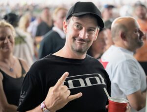 Smiling man in a black shirt and cap making a peace sign while holding a red cup with a straw.