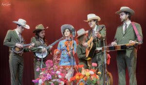 Band of six musicians in matching green pinstripe suits and cowboy hats on stage, playing guitars, violin, banjo, with colorful flowers in front.