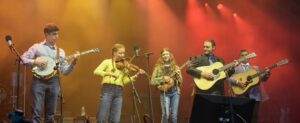 Group of five young musicians performing on a stage with guitars, a violin, and microphones under warm orange-red lighting.