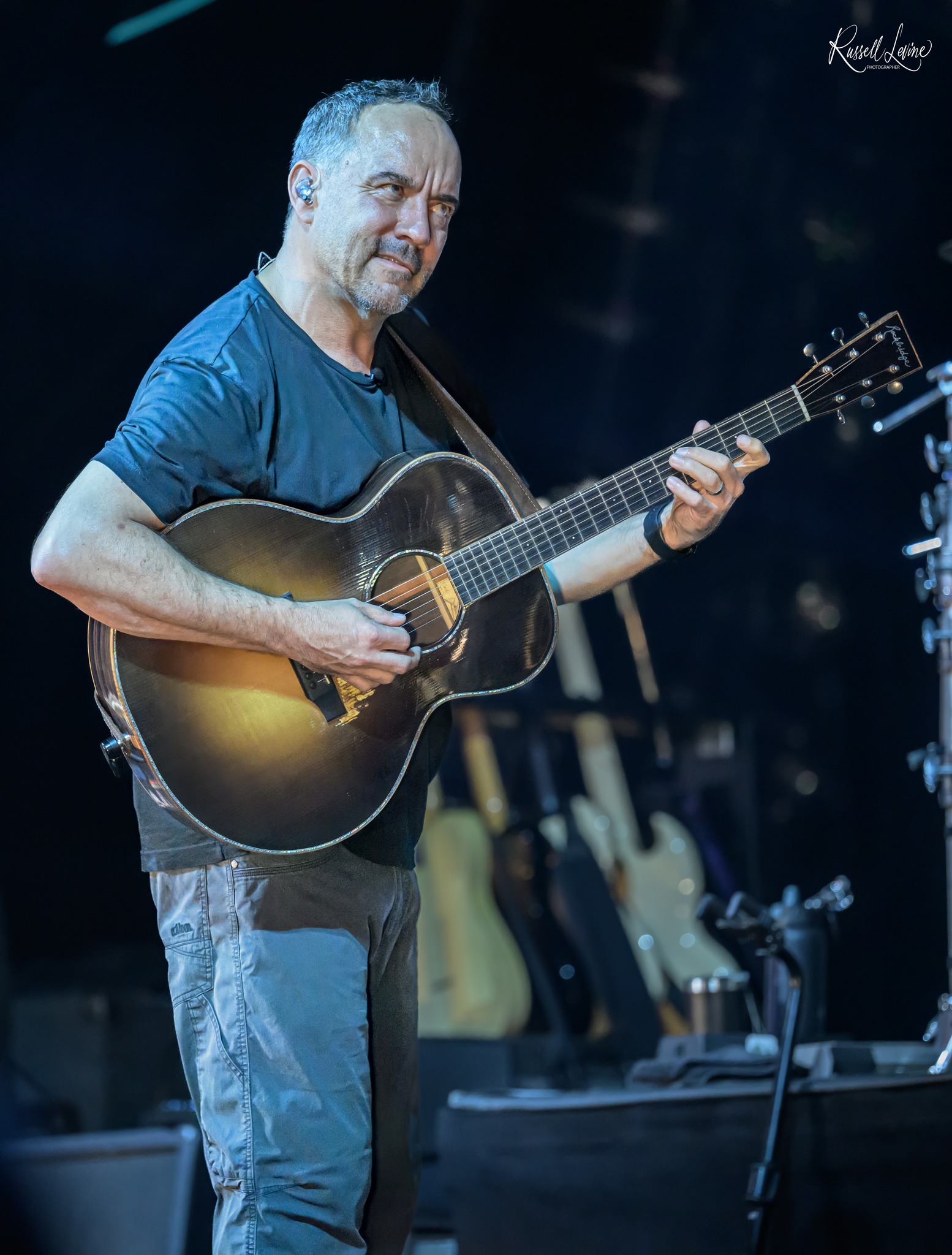 Male guitarist on a dim stage, playing a sunburst acoustic guitar with in-ear monitor in his ear.