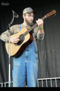 Bearded musician playing an acoustic guitar on stage, wearing a camo shirt, blue overalls, cap, and a green wristband near a microphone.