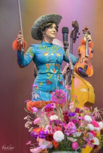 Female musician in a teal embroidered dress and large starry hat, holding a violin and bow at a microphone on a colorful stage, with pink flowers in the foreground.