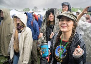 Smiling woman in a camo cap and rain jacket holding a blue festival cup at a rainy outdoor event, with others in ponchos in the background.